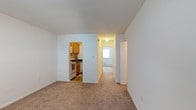 This single-floor residential unit offers a straightforward and functional layout designed for comfortable everyday living. The main living area is finished with wall-to-wall carpet, providing a warm and quiet underfoot feel, while the clean white walls throughout create a bright and neutral backdrop that suits a variety of décor styles. An open pass-through cutout connects the living space to the kitchen, which features warm wood-toned cabinetry, a dark countertop, and a freestanding gas range. The kitchen is finished with hard flooring, offering a practical contrast to the carpeted living area. A hallway extends from the main living space, leading toward additional rooms at the rear of the unit. Natural light filters in through a window visible at the end of the hallway, contributing to an airy feel throughout. A baseboard heating unit is visible along the hallway wall, indicating a reliable in-unit heating system. A door adjacent to the hallway entrance provides access to additional storage or a closet. Overall, the space presents a clean, unpretentious layout with practical finishes suited for functional residential use.