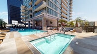 Located at 702 Broadway in the heart of downtown San Diego, this outdoor amenity level delivers a resort-caliber experience within an urban high-rise setting. The centerpiece of the space is a generously sized swimming pool with crystal-clear water, complemented by an adjacent spa offering a dedicated relaxation area. The surrounding sun deck is lined with cushioned lounge chairs, providing ample space for sunbathing beneath San Diego's abundant natural light.

A covered outdoor lounge area features a slatted pergola structure that provides shade while maintaining an open-air ambiance. Plush blue sofas, armchairs, and a wall-mounted television make this an ideal setting for both social gatherings and quiet relaxation. Additional seating vignettes are thoughtfully arranged throughout the deck, including intimate dining tables positioned to take full advantage of striking panoramic views of the downtown San Diego skyline, including landmark historic and modern high-rise buildings.

Lush palm trees and curated planters with ornamental grasses add natural texture and a California-inspired aesthetic to the space. The deck's clean, large-format concrete pavers and contemporary architectural detailing reflect the sleek, modern design of the surrounding tower. The open sightlines to the city create an immersive urban outdoor experience throughout the entire amenity level.