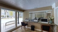 This single-floor office space is designed to serve as a professional and welcoming business or leasing center. The focal point of the room is a granite-topped service counter that spans a generous length, providing ample workspace and equipped with computers, a multifunction printer, and a monitor — ideal for administrative tasks and resident or client interactions. Behind the counter, horizontal blinds on the windows offer adjustable light control while maintaining privacy.

The space is finished with warm beige walls and durable tile flooring, creating a clean and practical environment. A textured ceiling adds a classic touch overhead, while a visible HVAC vent ensures comfortable climate control year-round. Seating is available along the counter, with cushioned chairs positioned for use by visitors or staff.

One of the standout features of this space is the large sliding glass window and door along one wall, which floods the room with natural light and offers a direct view of an outdoor pool area, complete with lounge furniture and surrounding fencing. This connection to the outdoor amenity space enhances the overall ambiance, giving the office an open, airy feel. The layout is efficiently organized to support day-to-day operations in a clear and accessible manner.