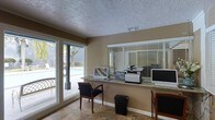 This single-floor office space is designed to serve as a professional and welcoming business or leasing center. The focal point of the room is a granite-topped service counter that spans a generous length, providing ample workspace and equipped with computers, a multifunction printer, and a monitor — ideal for administrative tasks and resident or client interactions. Behind the counter, horizontal blinds on the windows offer adjustable light control while maintaining privacy.

The space is finished with warm beige walls and durable tile flooring, creating a clean and practical environment. A textured ceiling adds a classic touch overhead, while a visible HVAC vent ensures comfortable climate control year-round. Seating is available along the counter, with cushioned chairs positioned for use by visitors or staff.

One of the standout features of this space is the large sliding glass window and door along one wall, which floods the room with natural light and offers a direct view of an outdoor pool area, complete with lounge furniture and surrounding fencing. This connection to the outdoor amenity space enhances the overall ambiance, giving the office an open, airy feel. The layout is efficiently organized to support day-to-day operations in a clear and accessible manner.