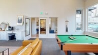 Located at 2920 Falk Rd in Vancouver, WA, this single-floor amenity space is thoughtfully designed to blend social, recreational, and fitness functions into one cohesive environment. Two entrances provide convenient access, welcoming visitors into a bright, airy lounge area distinguished by soaring ceilings, large windows that flood the space with natural light, and warm-toned hardwood-style flooring. A contemporary chandelier anchors the entry lounge, which features comfortable seating and a relaxed, modern aesthetic. The main lounge area continues the open, airy feel with high clerestory windows, warm leather seating, a wall-mounted television, and a natural wood media console. Adjacent to the lounge, a dedicated recreation area houses a full-sized billiards table, accented by large windows offering outdoor views and framed nature-inspired artwork, creating an inviting atmosphere for casual gatherings. The fitness center is comprehensively equipped with multiple treadmills, a cable strength machine, a full dumbbell rack, medicine balls, kettlebells, stability balls, and an Xponential+ interactive fitness station. Durable rubberized flooring and a large wall mirror complete the gym, which benefits from recessed lighting and vaulted ceilings. Together, these spaces offer a well-rounded, modern amenity experience.