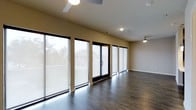 Located in Dallas, this single-floor space offers a clean, modern aesthetic defined by thoughtful design and quality finishes. The open layout is anchored by rich, dark hardwood flooring that runs throughout, providing a sleek and cohesive look underfoot. A striking wall of floor-to-ceiling windows with black framing floods the interior with abundant natural light, creating a bright and airy atmosphere while offering views of the surrounding neighborhood. Solar shades on the windows allow for flexible light control while maintaining the contemporary feel of the space. A sliding glass door provides direct access to the exterior, seamlessly connecting the indoors with the outside. The neutral beige walls complement the dark flooring and window frames, creating a balanced and inviting palette. Ceiling fans are positioned throughout to ensure comfortable airflow, and recessed lighting supplements the natural light for well-distributed illumination. The overall ambiance is modern and minimalist, making this Dallas space well-suited for a variety of uses, with its open plan offering versatility and a sense of spaciousness.
