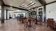 Located at 225 Sunlight Ln in Lake Mary, Florida, this single-floor community clubhouse offers a generously sized, open-plan gathering space designed for both relaxed socializing and casual entertaining. The interior is defined by a sophisticated blend of dark wood trim, coffered and tray ceilings with recessed lighting, and rich hardwood-style flooring laid in an eye-catching herringbone pattern throughout.

The main gathering area is anchored by a stone-surround fireplace with a wall-mounted television above, flanked by built-in display shelving. Bar-height round tables with leather-upholstered stools provide ample seating for groups. A dedicated lounge zone features plush upholstered armchairs and sofas arranged around a coffee table, bathed in natural light from large black-framed windows that frame views of the surrounding greenery.

A full kitchenette with dark cabinetry, granite countertops, a mosaic tile-fronted bar counter, stainless steel appliances, and pendant lighting serves as a functional hub for events. A quieter seating nook with a round table and patterned accent chairs offers an intimate conversation area. Additional flexible seating with ottomans and accent chairs rounds out the space, creating a warm, inviting atmosphere ideal for community gatherings.