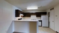This single-floor space is designed with practicality and functionality in mind. The kitchen serves as a central feature, showcasing dark espresso-toned cabinetry that lines the walls and provides generous storage. A peninsula counter extends outward, creating a natural breakfast bar that separates the kitchen from the adjacent living area while offering additional workspace. The countertops are finished in a clean white surface, complementing the white appliances, which include a full-size refrigerator and dishwasher. A double-basin sink is positioned along the back wall beneath a tiled backsplash, adding a clean, utilitarian aesthetic. An under-cabinet range hood is mounted above the cooktop area, ensuring proper ventilation. The kitchen is illuminated by a flush-mounted fluorescent light fixture, providing bright, even lighting throughout the space. The flooring transitions from light ceramic tile in the kitchen to carpet in the neighboring living area, delineating the two zones clearly. The textured ceiling adds a subtle dimensional quality to the interior. Overall, the space presents a clean, no-frills environment well-suited for everyday living.