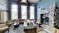 This single-floor space presents a striking interior defined by soaring double-height ceilings that lend an immediate sense of grandeur upon entry. The ceiling itself is a design feature, showcasing a geometric coffered detail finished in rich dark wood, from which stylish lantern-style pendant lights hang to provide warm, focused illumination alongside recessed lighting throughout.

The lounge area is anchored by an expansive arrangement of neutral-toned seating, including a large tufted sectional sofa accented with olive green throw pillows, complemented by tan accent chairs and upholstered ottomans. A round dark wood coffee table and stepped wooden side tables add refined contrast to the soft furnishings.

A wall of floor-to-ceiling windows dressed in flowing sheer drapes floods the space with abundant natural light while framing views of lush greenery outside. The walls are finished in a soothing slate blue tone that pairs harmoniously with the warm wood and neutral upholstery throughout.

A standout feature is the full-height tiled fireplace surround on one wall, clad in a mosaic of earth-toned tiles that adds texture and visual interest. Patterned carpeting grounds the seating area, contributing to the overall layered and cohesive design aesthetic. The space balances comfort and sophistication, making it ideal for both relaxed gatherings and quiet retreat.