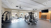Located at 1200 Trillium Cir in Raleigh, NC, this fitness center offers a generously sized, single-floor workout facility designed to accommodate a wide range of exercise needs. The space is thoughtfully laid out, with distinct zones for cardio, strength training, and free weights, allowing for an organized and efficient workout experience.

The main workout area features durable rubber flooring throughout, providing comfort and safety underfoot. A multi-station cable machine serves as the centerpiece of the strength training zone, complemented by a power rack and adjustable bench for functional lifting. The free weight area includes a full rack of dumbbells and a punching bag, set against a warm accent wall that adds a pop of color to the otherwise neutral interior. Cardio equipment includes multiple treadmills, a rowing machine, and a stationary bike, positioned near large windows that flood the space with natural light and offer pleasant outdoor views.

The ceiling is a standout architectural feature, with coffered detailing and multiple ceiling fans that ensure airflow and comfort during intense workouts. Recessed lighting throughout provides bright, even illumination. Wall-mounted mirrors in key areas enhance the sense of openness. The entrance leads seamlessly into the facility, and instructional signage is posted for user guidance. The overall ambiance is clean, modern, and welcoming.