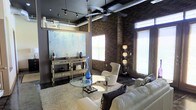 This single-floor space in Richardson, Texas, showcases a compelling blend of industrial and contemporary design elements. The living area immediately draws attention with its dramatic exposed brick walls that rise to soaring ceilings, lending the space a raw, urban character balanced by refined modern touches.

Overhead, exposed ductwork runs along the dark ceiling, reinforcing the industrial aesthetic, while a ceiling fan with a sleek profile provides both function and visual interest. Track lighting supplements the natural light that streams in through multiple large windows fitted with horizontal blinds, illuminating the space effectively throughout the day.

The living area is thoughtfully arranged with a plush white sofa and a sculptural white accent chair centered around a circular glass coffee table atop a soft area rug. A mirrored console table along one wall serves as an elegant display surface beneath a large-scale abstract artwork in cool blue and white tones. A tall floor lamp with stacked spherical shades adds ambient warmth to the corner.

The combination of polished concrete-style flooring, rich brick, dark ceiling, and curated furnishings creates a sophisticated, gallery-like ambiance that is both stylish and inviting.