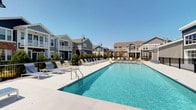 This residential community showcases a well-appointed outdoor amenity area centered around a generously sized swimming pool with crystal-clear blue water. The pool deck is finished in light-colored concrete and lined with a row of sling-style lounge chairs and side tables, offering ample space for sunbathing and relaxation. Black metal fencing borders the perimeter, providing both safety and a clean architectural boundary. Decorative street-style lamp posts are positioned throughout the deck, adding a charming, neighborhood aesthetic.

The surrounding buildings feature a contemporary design with a mix of gray and neutral-toned siding, pitched rooflines, and balconies with white railings, contributing to a cohesive and attractive streetscape. The multi-story structures overlook the pool area, creating an enclosed, community-oriented atmosphere. Under a clear blue sky, the outdoor space benefits from abundant natural light throughout the day, enhancing its open and inviting character. The overall design reflects a modern residential community with a strong emphasis on outdoor leisure and shared amenity spaces.