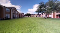 This single-floor residence is part of a well-maintained multi-building community set against a backdrop of lush, manicured green lawns and mature trees, creating a welcoming and open outdoor environment. The exteriors of the buildings feature warm earth-tone siding and traditional architectural styling, contributing to a cohesive and inviting community aesthetic.

Inside, the unit showcases a thoughtfully designed interior that blends industrial and contemporary elements. The dining area is a standout space, anchored by a striking round green marble table paired with woven metal chairs, all set atop a natural fiber area rug. Polished concrete floors run throughout, lending the interior an urban, modern character while remaining highly durable.

The walls are finished in a clean, neutral off-white palette that provides an ideal backdrop for curated artwork, including a large abstract framed piece that adds a refined, gallery-like quality to the dining space. A sleek sliding barn door with black metal hardware serves as both a functional and decorative divider, enhancing the modern-industrial atmosphere.

Recessed lighting provides focused illumination throughout, while the open sightlines between rooms create a sense of flow and connectivity. Transitional doorways with aged wood detailing add warmth and character, balancing the cooler concrete and metal finishes found throughout the space.