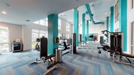 Located at 215 107th Street Circle East in Bradenton, Florida, this single-floor fitness center is a well-appointed, contemporary facility designed to support a comprehensive workout experience. The space is immediately striking upon entry, with glass-paneled doors opening into a bright, airy interior that exudes a clean, modern aesthetic.

The main workout floor is defined by soaring double-height ceilings and bold teal-painted columns that rise dramatically throughout the space, lending a vibrant, energetic atmosphere. Large windows line the walls on multiple sides, flooding the interior with natural light and offering views of the surrounding Florida landscape, including palm trees and lush greenery. A mezzanine-level overlook with glass railings adds to the architectural depth of the room.

The fitness floor is outfitted with a wide variety of equipment, including treadmills, stationary bikes, ellipticals, cable machines, free-weight stations with a full dumbbell rack, and multiple strength training machines — accommodating both cardio and resistance training. Full-length mirrors along one wall enhance the sense of openness while providing functional workout visibility.

Underfoot, a stylish herringbone-patterned carpet in blue and neutral tones adds a polished design touch throughout. A quieter corner area features floor-to-ceiling windows, a beverage refrigerator, and open shelving, offering a welcoming cooldown zone. The overall ambiance is bright, motivating, and meticulously maintained.