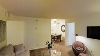 This single-story residence presents an open, connected layout between the living and dining areas, creating a cohesive and functional living environment. The living area features warm-toned carpeting underfoot and neutral cream-colored walls that lend a clean, understated aesthetic. A glass-topped coffee table and seating arrangement anchor the space, while a decorative floral arrangement adds a touch of life to the room.

The adjoining dining area is visible through an open passageway, maintaining a sense of flow between the two spaces. It is equipped with a ceiling fan fitted with a light fixture, providing both ventilation and overhead illumination. A round dining table with chairs sits beneath this fixture, and a circular decorative mirror mounted on the wall adds visual interest to the dining space.

Consistent carpeting and matching wall tones run throughout both areas, reinforcing the unified design language of the home. A nearby door provides access to additional spaces within the residence. The overall ambiance is warm and comfortable, with a straightforward layout suited for everyday living and casual dining.