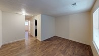 This single-story space is designed with practicality and comfort in mind, showcasing a cohesive aesthetic carried throughout each room by warm, medium-toned hardwood-style flooring and crisp white walls with clean white baseboards. The main living area is open and well-proportioned, receiving natural light through a window fitted with blinds, and features a front entry door. The layout connects seamlessly to additional living space, visible through an open passage that also provides a glimpse into the kitchen beyond.

The galley-style kitchen is efficiently arranged, featuring dark espresso cabinetry both above and below the counters, paired with granite-patterned countertops that lend a polished look. A double stainless steel sink, electric range, refrigerator, and over-range microwave complete the functional setup, all finished in a coordinated black appliance suite.

The bedroom is a comfortable, well-lit room with a ceiling fan with light fixture and a window with blinds providing natural light and ventilation. The bathroom is finished with a clean, white soaking tub and shower combination equipped with built-in shelving and a chrome shower rod and fixtures, maintaining the unit's simple and tidy overall character.
