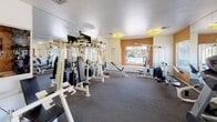 This single-floor fitness center offers a dedicated space for strength training and exercise. The room is furnished with an extensive selection of weight machines and cable equipment arranged throughout the space, providing users with options for a full-body workout. The flooring consists of durable rubber matting, ideal for a high-traffic gym environment and providing both safety and stability during exercise.

Mirrored walls line one side of the room, visually expanding the space while allowing users to monitor their form during workouts. The ceiling features a textured finish and is equipped with circular overhead light fixtures that provide consistent, even illumination throughout the area. A warm accent wall in a muted orange tone adds a subtle visual contrast to the otherwise neutral color palette of white walls and grey flooring.

A window on the far wall allows natural light to enter the space and offers a view of the surrounding exterior, contributing to an open and airy atmosphere. The overall layout is practical and functional, with equipment arranged to maximize usable floor space while maintaining comfortable circulation throughout the fitness center.
