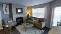 This single-floor residence offers a thoughtfully designed living space that balances comfort and style. The living room serves as the heart of the home, anchored by a tiled fireplace framed by a decorative mirror, providing a warm and inviting focal point. Neutral gray and beige wall tones create a calming backdrop that complements the warm-toned sofa, which is dressed with navy and patterned throw pillows for a layered, curated look. A large window dressed with white blinds and sheer curtains allows generous natural light to fill the room, enhancing the airy feel of the space. Dark hardwood flooring grounds the room and adds a sense of warmth and sophistication. A round wooden coffee table with hairpin legs sits atop a chevron-patterned area rug, adding a contemporary touch to the overall design. Ambient lighting from a tripod floor lamp contributes to the cozy atmosphere during evening hours. Subtle decorative accents, including a gold leaf wall art piece and natural elements on the coffee table, reflect an attention to detail throughout. The space presents a seamless blend of modern and transitional design elements, making it both functional and visually appealing.