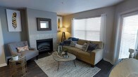 This single-floor residence offers a thoughtfully designed living space that balances comfort and style. The living room serves as the heart of the home, anchored by a tiled fireplace framed by a decorative mirror, providing a warm and inviting focal point. Neutral gray and beige wall tones create a calming backdrop that complements the warm-toned sofa, which is dressed with navy and patterned throw pillows for a layered, curated look. A large window dressed with white blinds and sheer curtains allows generous natural light to fill the room, enhancing the airy feel of the space. Dark hardwood flooring grounds the room and adds a sense of warmth and sophistication. A round wooden coffee table with hairpin legs sits atop a chevron-patterned area rug, adding a contemporary touch to the overall design. Ambient lighting from a tripod floor lamp contributes to the cozy atmosphere during evening hours. Subtle decorative accents, including a gold leaf wall art piece and natural elements on the coffee table, reflect an attention to detail throughout. The space presents a seamless blend of modern and transitional design elements, making it both functional and visually appealing.