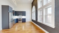 This thoughtfully designed 2-bedroom, 2-bathroom residence seamlessly blends historic architectural character with contemporary finishes across a single floor. The open kitchen and living area serves as the heart of the home, distinguished by dramatically high ceilings, warm honey-toned hardwood floors, and a striking feature wall of exposed dark brick framing a series of large arched and rectangular windows that flood the space with natural light and offer views of the surrounding urban streetscape. The kitchen is outfitted with sleek slate-blue cabinetry, stainless steel appliances including a range, microwave, refrigerator, and dishwasher, complemented by white quartz countertops and reclaimed wood open shelving that add a rustic-modern contrast. Both bedrooms are bright and well-proportioned, featuring the same warm hardwood flooring and clean white walls. The primary bedroom includes a double-door closet and soaring ceilings. The bathrooms are finished with large-format stone tile flooring, floating wood vanities with quartz surfaces, black fixtures, and white subway tile. A hallway and utility room round out the functional layout, making this a well-appointed and characterful residence.