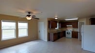 This single-story home offers a practical and open layout, seamlessly connecting the living area and kitchen into one cohesive space. The living area is finished with light-toned tile flooring that extends throughout, creating a clean and uniform look. A ceiling fan with a built-in light fixture provides both comfort and illumination, while a window along the exterior wall allows natural light to fill the space. The kitchen is well-appointed with rich, dark wood cabinetry that provides ample storage through both upper and lower cabinet configurations. A double-basin sink is positioned beneath a window, offering a pleasant view while working. The kitchen is equipped with a white range and overhead ventilation hood, complemented by under-cabinet lighting above the cooking area. Dark granite-style countertops contrast nicely with the warm wood cabinetry, adding a touch of character to the space. A dishwasher is integrated into the lower cabinetry, completing the functional kitchen setup. The neutral wall tones throughout lend a warm and inviting ambiance, while the open sightlines between the living and kitchen areas make the overall space feel airy and versatile.