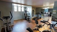 This single-floor fitness space is thoughtfully designed to support a well-rounded exercise routine. The room features warm hardwood-style flooring that extends throughout, providing both durability and a clean, polished aesthetic. The walls are painted in a cool gray tone, contributing to a focused and energizing atmosphere.

The space is outfitted with a range of exercise equipment, including a multi-function cable and weight machine, two treadmills, and a recumbent bike, offering options for both strength training and cardiovascular workouts. A large wall-mounted mirror on one side of the room enhances the sense of space and allows users to monitor their form during exercise.

Natural light filters in through multiple windows on one wall, brightening the room and creating an open, airy feel. A ceiling fan is centrally positioned to provide ventilation and comfort during intense workouts. An interior door provides access to an adjacent area, adding to the overall functionality of the layout.

The textured ceiling and clean wall finishes reflect a practical, no-frills design approach suited to a dedicated fitness environment. Overall, this space delivers a compact yet comprehensive gym experience with a comfortable and well-lit setting.