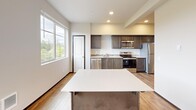 Located at 102 E Division St in Arlington, Washington, this single-floor unit offers a thoughtfully designed open layout making efficient use of its space. The kitchen serves as the central feature of the unit, showcasing dark espresso-toned cabinetry paired with light quartz countertops for a clean, contemporary contrast. A full suite of stainless steel appliances includes an electric range, built-in microwave, dishwasher, and refrigerator, providing complete functionality. A kitchen island with matching cabinetry and a white countertop offers additional prep and storage space while visually anchoring the open-concept layout.

Light wood-tone flooring runs throughout, lending warmth and continuity to the interior. Recessed lighting illuminates the space evenly, complemented by large windows that draw in natural light and frame views of the surrounding Arlington landscape, including glimpses of the nearby mountains. The neutral wall tones and clean architectural lines contribute to a bright, modern ambiance. The open area adjacent to the kitchen provides flexible living space, completing this efficiently designed, move-in-ready unit.