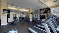 This single-level fitness center offers a comprehensive workout environment designed to accommodate a variety of exercise routines. The space is outfitted with a robust selection of cardio machines, including treadmills and stationary bikes positioned toward the front, alongside elliptical trainers and rowing equipment spread throughout the floor. A dedicated strength training area features cable machines, weight benches, and free weights, providing options for full-body conditioning.

The flooring consists of durable dark rubber matting, ideal for both safety and equipment stability. Mirrored walls line one side of the room, enhancing the sense of space while allowing users to monitor their form during workouts. Multiple wall-mounted flat-screen televisions are strategically placed for visibility from various workout stations. The ceiling is finished with recessed and track lighting that maintains a bright, functional atmosphere. Motivational fitness artwork and informational charts adorn the walls, adding character to the space. Large windows near the entrance allow natural light to filter in, contributing to an open and energizing environment.