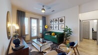 This single-floor residence combines contemporary design with thoughtful functionality across a well-appointed layout. The living room serves as the centerpiece, featuring hardwood-style flooring, a ceiling fan, and large windows with blinds and draping curtains that invite abundant natural light. A gallery wall of framed black-and-white photography adds artistic character to the space.

The kitchen is outfitted with stainless steel appliances, two-tone cabinetry pairing light upper and dark lower cabinets, a subway tile backsplash, and a substantial island with dark granite countertops and pendant lighting overhead — providing both prep space and casual seating.

Two bedrooms offer carpeted flooring and large windows. The primary bedroom provides a spacious, tranquil retreat, while a secondary room functions effectively as a home office, complete with a glass-top desk positioned to take advantage of natural light.

Both bathrooms feature wood-look flooring, dark granite vanities, and grey cabinetry. One includes a glass-enclosed walk-in shower with marble-style tile and access to a walk-in closet, while the other offers a tub-shower combination and an adjacent closet for additional storage.