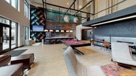 Located at 6847 Stewart Rd in Cincinnati, Ohio, this single-floor clubhouse facility is designed with a contemporary aesthetic that balances sophistication and social functionality. The entry greets visitors with a sleek reception desk finished in white veined stone, set beneath a slatted wood ceiling accented by linear LED lighting — creating a warm yet polished first impression with views toward the exterior entrance.

The main lounge area features ambient cove lighting, plush seating arrangements including navy blue sofas, leather accent chairs, and upholstered ottomans atop textured carpeting. Artistic branch-patterned wall treatments in deep charcoal add a distinctive design element throughout the space.

A dramatic double-height games and social lounge serves as a central focal point, anchored by a tournament-style billiards table beneath globe pendant lights suspended from an exposed industrial ceiling grid. A full wet bar with counter seating, a large-format mural wall in rich patina tones, and tufted wine-colored accent panels contribute to the vibrant, loft-inspired atmosphere.

A separate seating alcove offers rich leather sectional sofas alongside floor-to-ceiling windows that frame direct views of a landscaped courtyard with outdoor seating, string lights, and the surrounding residential buildings — seamlessly connecting the interior to the outdoor amenity spaces.