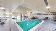 Located at 15 Iron Horse Dr in Bedford, New Hampshire, this single-floor indoor aquatic facility offers a well-appointed and inviting environment for swimming and relaxation. The centerpiece of the space is a generously sized indoor lap pool with crystal-clear turquoise water, lane markings, and underwater lighting that creates a bright, welcoming atmosphere. The pool is surrounded by a wide tiled deck furnished with lounge chairs and small tables, providing ample space for relaxation before or after a swim.

The facility's architecture is defined by a dramatic vaulted ceiling with clean white paneling that rises to a substantial peak, lending the space an open, airy feel. Large windows line the perimeter walls, flooding the interior with natural light and offering views of the surrounding landscape. A decorative diamond-patterned tile border runs along the walls, adding a classic, polished aesthetic to the space.

Separate from the main pool area, a dedicated spa room houses an octagonal hot tub with tiered seating, tiled in a coordinating diamond motif. Posted spa rules and thoughtful lighting make this a defined, comfortable retreat. Together, the pool and spa areas create a comprehensive aquatic experience in a bright, well-maintained, and architecturally refined setting.
