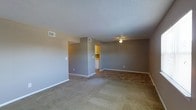 This single-floor residential space offers a clean, functional layout designed for everyday comfort. The main living area is generously proportioned, featuring soft wall-to-wall carpeting in a neutral tone that adds warmth underfoot. The walls are finished in a contemporary gray palette, complemented by crisp white baseboards that provide a clean visual contrast throughout. A ceiling fan is centrally mounted, ensuring comfortable airflow across the living space.

The textured popcorn ceiling adds a classic residential character, while multiple HVAC vents are strategically positioned to maintain consistent climate control. The open layout connects seamlessly to an adjacent area, which transitions into the kitchen. The kitchen is visible through a doorway and features warm wood-toned cabinetry and tile flooring, offering a functional cooking and storage space. White appliances are integrated into the kitchen, maintaining a tidy and cohesive aesthetic.

The overall ambiance of the space is practical and move-in ready, with a straightforward design that prioritizes usability. Neutral tones and simple finishes create a versatile backdrop easily adaptable to a variety of interior styles. The single-floor layout ensures easy accessibility and flow between spaces, making this a well-organized and efficiently designed residential unit.