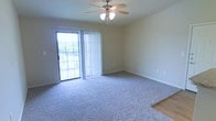 This single-floor space features a well-proportioned living area finished with plush wall-to-wall carpeting in a neutral gray tone, providing a comfortable and understated foundation. The room transitions to hard flooring at the edges, visible near the entryway, adding a subtle contrast in materials. A ceiling fan with an integrated light fixture is centrally mounted, offering both air circulation and ambient lighting throughout the space.

A sliding glass door dominates one wall, fitted with vertical blinds that allow for adjustable light control and privacy. The door opens to an outdoor balcony, visible through the glass, where greenery and an open view can be seen — bringing a sense of connection to the outdoors. This feature allows natural light to fill the room during the day, contributing to a bright and airy atmosphere.

The walls are finished in a soft, warm beige tone that complements the neutral carpeting, while white baseboards provide clean visual definition. Electrical outlets are positioned conveniently along the walls. Overall, the space presents a functional, move-in-ready layout with a calm and practical aesthetic suited to a variety of uses.
