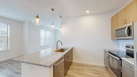 This well-appointed single-floor unit offers a clean, modern aesthetic defined by a consistent palette of crisp white walls, light wood-look flooring, and slate-gray interior doors with black hardware. The open-concept kitchen and living area serve as the heart of the space, where a granite-topped island with an undermount brass sink and stainless steel dishwasher anchors the layout. Warm pendant lighting with globe fixtures adds character overhead, while light wood cabinetry, a subway tile backsplash, and a full suite of stainless steel appliances — including a refrigerator, electric range, and built-in microwave — complete the kitchen. The adjacent living area is generously proportioned, bathed in natural light from multiple windows and a set of French doors that open to an exterior space. The bedrooms feature plush carpeting, ceiling fans, and large windows that bring in ample daylight. The bathrooms are finished with granite-topped vanities, tile walk-in showers, and light hardwood-style flooring. A hallway with recessed lighting and a transom-vented closet connects the rooms with functional ease, contributing to an overall ambiance that is bright, contemporary, and move-in ready.