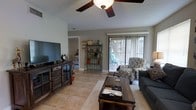 This single-floor residence offers a welcoming and functional layout designed for comfortable everyday living. The living room serves as the heart of the home, featuring large-format tile flooring that extends throughout the space, providing a clean and cohesive look that is both practical and visually appealing. A ceiling fan with a light fixture is centrally positioned overhead, ensuring year-round comfort and efficient air circulation.

Natural light fills the room through a sliding glass door fitted with vertical blinds, which opens directly to an outdoor patio area — ideal for enjoying fresh air and outdoor relaxation. The living area is thoughtfully arranged to accommodate a large flat-screen television set on a dark wood media console, complemented by a matching coffee table that anchors the seating arrangement. A decorative metal shelving unit adds a charming, personalized touch to the space.

A textured ceiling adds subtle visual character, while neutral-toned walls create a versatile backdrop that complements a range of furnishing styles. An open sightline connects the living room to adjacent areas of the home, reinforcing the spacious and airy feel of the layout. Overall, this residence combines practical design with warm, lived-in character.