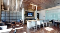 This conference room offers a sophisticated blend of modern design and practical functionality. The space is immediately distinguished by its striking wood-planked ceiling, which adds warmth and texture while complementing the rich hardwood flooring below. A recessed tray detail in the ceiling frames the room's central gathering area and houses sleek pendant lighting that provides focused illumination.

At the heart of the room sits a substantial island with a polished granite countertop, surrounded by contemporary upholstered chairs in a neutral gray tone — ideal for informal meetings or collaborative work sessions. A wall-mounted flat-screen TV anchors the media wall, which features a dark, textured accent panel with integrated ambient backlighting, creating a refined focal point for presentations or viewing.

Along one wall, a generously proportioned upholstered bench with tall, padded gray panels provides additional seating, accompanied by sculptural wooden accent chairs for a more relaxed conversation area. The combination of natural materials, carefully layered lighting, and clean-lined furnishings gives the space a polished, professional ambiance that remains warm and inviting.