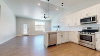 This two-story home in Herriman, Utah offers four bedrooms and three bathrooms across a generously sized floor plan designed for comfortable, modern living. The main level centers on an open-concept kitchen and living area distinguished by soaring vaulted ceilings that amplify the sense of space and airiness. The kitchen is outfitted with white shaker-style cabinetry, matte black hardware, light quartz countertops, and stainless steel appliances including a gas range, over-the-range microwave, and dishwasher. A peninsula provides additional prep and seating space, while pendant lighting adds a contemporary accent overhead. Recessed lighting and a ceiling fan with a light kit serve the adjoining living area, which receives natural light through a large window with outdoor views. Durable wood-look flooring runs throughout the main living spaces, lending warmth and continuity. The bedrooms are carpeted and well-lit with natural light from windows. A primary bathroom features a dual-sink vanity with white cabinetry, matte black fixtures, a large mirror, industrial-style sconce lighting, and elegant tile flooring. The primary suite also includes a walk-in closet with built-in hanging rods. The home's clean, neutral palette and consistent design language create a cohesive, move-in-ready aesthetic throughout.