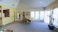 This single-floor space offers a welcoming and airy atmosphere, defined by its striking vaulted ceilings that amplify the sense of openness throughout the interior. The main area is finished with soft, wall-to-wall carpeting in a neutral gray tone, providing a comfortable and cohesive foundation for the room.

A central dining arrangement anchors the space, featuring a round wooden table surrounded by cross-back chairs, creating an inviting setting for gatherings. A framed nature-themed artwork adorns one of the walls, adding a subtle decorative touch to the otherwise clean, minimalist aesthetic.

Natural light is a defining characteristic of this space, with a generous row of large windows along one wall that frame views of mature greenery outside. The windows flood the interior with daylight and create a pleasant connection to the outdoors.

A set of classic French doors with glass panes opens to an adjacent room, adding architectural charm and visual depth to the layout. Additional doorways provide access to connected spaces, suggesting a functional and well-connected floor plan. Recessed lighting in the ceiling complements the abundant natural light, ensuring the space remains bright and welcoming throughout the day. The overall ambiance is clean, open, and versatile.