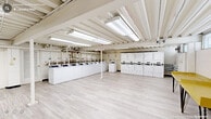 Located at 2517 Guess Rd in Durham, NC, this single-floor community utility facility offers a generous, open layout designed to serve the practical needs of its users. The space is thoughtfully organized into two primary functional zones: a full-scale laundry area and a dedicated mailroom section.

The laundry area is the centerpiece of the facility, equipped with an extensive row of commercial-grade top-loading washing machines and a matching bank of dryers. The machines are arranged along the perimeter walls, leaving a wide, open floor area that provides comfortable circulation and accommodates folding tables for added convenience. A yellow utility table is also present for resident use.

The mailroom section houses multiple rows of individual tenant mailboxes in a tan finish, organized in multi-unit clusters and accessible with individual keys. A simple bench near the exit provides a resting spot for visitors.

Throughout the space, light-toned luxury vinyl plank flooring lends a clean, modern aesthetic. The exposed structural ceiling, painted white and fitted with bright fluorescent lighting panels, ensures the entire area is well-illuminated. Visible utility piping and conduit run along the ceiling in an organized manner. White-painted concrete block walls and clearly marked exit doors contribute to the functional, no-frills character of this well-maintained shared amenity space.