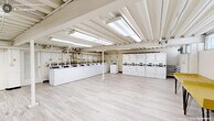 Located at 2517 Guess Rd in Durham, NC, this single-floor community utility facility offers a generous, open layout designed to serve the practical needs of its users. The space is thoughtfully organized into two primary functional zones: a full-scale laundry area and a dedicated mailroom section.

The laundry area is the centerpiece of the facility, equipped with an extensive row of commercial-grade top-loading washing machines and a matching bank of dryers. The machines are arranged along the perimeter walls, leaving a wide, open floor area that provides comfortable circulation and accommodates folding tables for added convenience. A yellow utility table is also present for resident use.

The mailroom section houses multiple rows of individual tenant mailboxes in a tan finish, organized in multi-unit clusters and accessible with individual keys. A simple bench near the exit provides a resting spot for visitors.

Throughout the space, light-toned luxury vinyl plank flooring lends a clean, modern aesthetic. The exposed structural ceiling, painted white and fitted with bright fluorescent lighting panels, ensures the entire area is well-illuminated. Visible utility piping and conduit run along the ceiling in an organized manner. White-painted concrete block walls and clearly marked exit doors contribute to the functional, no-frills character of this well-maintained shared amenity space.