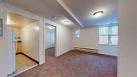 This ground-level unit offers a practical and comfortable layout spread across a single floor. The main living area is generously sized, featuring warm neutral carpeting, exposed structural beams on the ceiling, baseboard heating, and multiple windows that allow natural light to filter in, offering views of the surrounding brick exterior. The space flows seamlessly into a secondary room, ideal as a bedroom or flexible living space, which includes a large sliding-door closet and its own window.

A dedicated bedroom provides a quieter retreat with plush carpeting, a bright window with blinds, and a baseboard heater for year-round comfort. The kitchen is efficiently designed with light maple cabinetry, rich dark granite countertops, a white gas range with overhead ventilation, a corner sink, and durable tile flooring. A subway tile backsplash adds a clean, classic touch.

The full bathroom includes a vanity with white cabinetry, a toilet, and a tub-shower combination, all finished with neutral tile flooring and a window for ventilation. A warm wood-toned entry door welcomes residents into the carpeted foyer, setting the tone for the unit's clean, functional aesthetic throughout.
