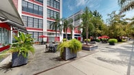 Located at 3700 Orleans Ave in New Orleans, this expansive outdoor space is defined by a bold architectural backdrop — a multi-story building clad in vivid red brick accented with white detailing and large industrial-style windows that flood the grounds with natural light. The courtyard is thoughtfully designed to accommodate a variety of uses, offering multiple distinct seating zones. Open-air dining areas are furnished with sleek patio tables and chairs, some shaded by red umbrellas, while modern high-top tables and contemporary stools line a covered structural canopy supported by steel lattice framework. A manicured turf lawn provides a relaxed lounge area outfitted with wicker furniture and colorful accent chairs, ideal for casual gatherings. Throughout the grounds, mature palm trees, cypress trees, and lush tropical plantings in large corrugated metal planters lend a distinctly New Orleans character to the space. Concrete walkways connect the various zones, bordered by well-tended garden beds. A gated archway framed by the red brick structure serves as a striking architectural focal point. The overall ambiance is lively yet welcoming, blending industrial heritage with verdant, tropical outdoor living in the heart of New Orleans.