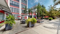 Located at 3700 Orleans Ave in New Orleans, this expansive outdoor space is defined by a bold architectural backdrop — a multi-story building clad in vivid red brick accented with white detailing and large industrial-style windows that flood the grounds with natural light. The courtyard is thoughtfully designed to accommodate a variety of uses, offering multiple distinct seating zones. Open-air dining areas are furnished with sleek patio tables and chairs, some shaded by red umbrellas, while modern high-top tables and contemporary stools line a covered structural canopy supported by steel lattice framework. A manicured turf lawn provides a relaxed lounge area outfitted with wicker furniture and colorful accent chairs, ideal for casual gatherings. Throughout the grounds, mature palm trees, cypress trees, and lush tropical plantings in large corrugated metal planters lend a distinctly New Orleans character to the space. Concrete walkways connect the various zones, bordered by well-tended garden beds. A gated archway framed by the red brick structure serves as a striking architectural focal point. The overall ambiance is lively yet welcoming, blending industrial heritage with verdant, tropical outdoor living in the heart of New Orleans.