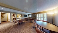 This single-floor community room offers a comfortable and functional setting designed for group gatherings and social interaction. The main lounge and dining area is generously sized, featuring multiple round tables paired with cushioned chairs arranged throughout the carpeted space. Textured ceilings with recessed lighting provide a warm, even illumination throughout the room, while large windows fitted with plantation shutters allow natural light to filter in and offer views of the surrounding greenery. Decorative wall art, including a metal tree sculpture, a sunflower print, a large clock, and floral accents, lends a welcoming character to the space. A wall-mounted flat-screen television serves as a focal point on one end of the room, making the area suitable for presentations or shared viewing. A wall unit provides supplemental climate control. Adjacent to the main gathering area is a compact but well-equipped kitchen, outfitted with white cabinetry, a full-size refrigerator, an electric range with overhead ventilation, and ample counter space. A small restroom with a pedestal sink rounds out the facility, ensuring full convenience for occupants and guests.