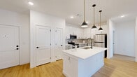 Located on N Marksheffel Rd in Colorado Springs, CO, this well-appointed one-bedroom, one-bathroom unit offers a thoughtfully designed single-level layout with modern finishes throughout. The open-concept kitchen and living area serves as the heart of the home, showcasing crisp white upper cabinetry, gray lower cabinets with a subtle hexagonal tile backsplash, and a full suite of stainless steel appliances including a refrigerator, range, and microwave. A large center island with a white quartz countertop and undermount sink is illuminated by a trio of statement black-and-gold pendant lights, providing both functionality and style. Warm-toned hardwood-style flooring flows seamlessly throughout the entire unit, complementing the bright white walls and recessed lighting. The living area extends toward a glass exterior door and large window, inviting abundant natural light and outdoor views. The bedroom is a comfortable, well-proportioned retreat featuring a ceiling fan, a natural light-filled window, and access to a dedicated closet. The bathroom is finished with a wood-look tile accent wall, a wide vanity with white quartz countertop, black matte fixtures, and a backlit LED mirror. An in-unit laundry area adds everyday convenience to this clean, contemporary Colorado Springs residence.