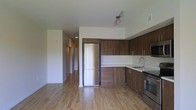 This single-floor space showcases a clean, contemporary design with an emphasis on functionality and modern aesthetics. The open-concept kitchen serves as a central highlight, outfitted with rich, warm-toned wood cabinetry that extends from floor to ceiling, providing generous storage. Stainless steel appliances — including a full-size refrigerator, stove, and built-in microwave — complement the crisp white countertops, creating a sleek contrast. A stainless steel sink is neatly integrated into the counter space, and track lighting above adds focused illumination to the work area. Light hardwood flooring runs seamlessly throughout the space, contributing to a bright and airy atmosphere. The open layout connects the kitchen to the adjacent living area, enhancing the sense of spaciousness. A hallway visible from the main area leads to additional rooms, hinting at a well-organized floor plan. The white walls and clean lines throughout maintain a minimalist character, making the space feel fresh, versatile, and move-in ready. Overall, the design balances practical living with a polished, modern aesthetic.