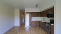 This single-floor space showcases a clean, contemporary design with an emphasis on functionality and modern aesthetics. The open-concept kitchen serves as a central highlight, outfitted with rich, warm-toned wood cabinetry that extends from floor to ceiling, providing generous storage. Stainless steel appliances — including a full-size refrigerator, stove, and built-in microwave — complement the crisp white countertops, creating a sleek contrast. A stainless steel sink is neatly integrated into the counter space, and track lighting above adds focused illumination to the work area. Light hardwood flooring runs seamlessly throughout the space, contributing to a bright and airy atmosphere. The open layout connects the kitchen to the adjacent living area, enhancing the sense of spaciousness. A hallway visible from the main area leads to additional rooms, hinting at a well-organized floor plan. The white walls and clean lines throughout maintain a minimalist character, making the space feel fresh, versatile, and move-in ready. Overall, the design balances practical living with a polished, modern aesthetic.