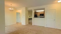 This single-floor space features an open and functional layout that connects the main living area to the kitchen through a half-wall pass-through design. The living area is finished with wall-to-wall beige carpeting, lending a warm and neutral tone throughout the space. Soft cream-colored walls enhance the brightness of the interior, creating an inviting atmosphere.

The kitchen is positioned at the rear of the unit and is accessible via the pass-through counter, which also serves as a visual divider between the cooking and living areas. Light wood cabinetry lines the upper and lower sections of the kitchen, offering ample storage. A black microwave and refrigerator provide modern appliances, while dark countertops contrast neatly against the lighter cabinetry. Recessed and overhead fluorescent lighting ensures the kitchen is well-lit.

A hallway visible from the main living area leads to additional rooms, suggesting a well-organized floor plan. A doorway at the end of the hall opens to what appears to be a secondary space, while another door on the right side of the living area indicates additional access points within the unit. The overall ambiance is clean, neutral, and move-in ready.