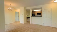 This single-floor space features an open and functional layout that connects the main living area to the kitchen through a half-wall pass-through design. The living area is finished with wall-to-wall beige carpeting, lending a warm and neutral tone throughout the space. Soft cream-colored walls enhance the brightness of the interior, creating an inviting atmosphere.

The kitchen is positioned at the rear of the unit and is accessible via the pass-through counter, which also serves as a visual divider between the cooking and living areas. Light wood cabinetry lines the upper and lower sections of the kitchen, offering ample storage. A black microwave and refrigerator provide modern appliances, while dark countertops contrast neatly against the lighter cabinetry. Recessed and overhead fluorescent lighting ensures the kitchen is well-lit.

A hallway visible from the main living area leads to additional rooms, suggesting a well-organized floor plan. A doorway at the end of the hall opens to what appears to be a secondary space, while another door on the right side of the living area indicates additional access points within the unit. The overall ambiance is clean, neutral, and move-in ready.