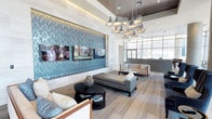 This contemporary lounge space showcases a thoughtfully designed interior that blends modern aesthetics with functional comfort. The most commanding feature is a large-scale teal mosaic tile accent wall, which serves as the room's visual centerpiece. Framed by clean white borders, the wall incorporates multiple flat-screen televisions, creating an engaging entertainment focal point.

The ceiling draws the eye upward with its layered design, incorporating subtle recessed lighting alongside a dramatic cluster of metallic pendant lights. These sculptural fixtures provide both ambient illumination and a striking decorative element that anchors the seating area below.

The lounge is furnished with a carefully curated collection of contemporary pieces, including a chaise lounge, upholstered sofas, and accent chairs in neutral tones of gray and beige. Dark accent tables provide contrast and balance to the lighter palette. Light wood-toned flooring extends throughout, complementing the overall color scheme.

Large floor-to-ceiling windows along one wall flood the space with natural light and offer views to the exterior, creating a seamless connection between the interior and the outside environment. The overall ambiance is polished and welcoming.