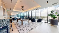 Located in the heart of Miami's vibrant Wynwood neighborhood, this upscale amenity lounge offers a generous, open-plan layout designed for both socializing and relaxation. The space is defined by an expansive curtain wall of floor-to-ceiling glass panels along its entire length, flooding the interior with abundant natural light and framing sweeping views of the Miami skyline and surrounding urban landscape. Large-format porcelain tile flooring runs throughout, anchored by a bold, abstract area rug that delineates the central gathering zone.

The living area is thoughtfully divided into distinct functional zones. A game area centers on a handsome billiard table beneath a dramatic multi-ring chandelier suspended from a coffered, cove-lit ceiling. Adjacent lounge seating clusters feature a mix of sculptural black accent chairs and plush cream armchairs arranged around a sleek stone coffee table, ideal for intimate conversation. A built-in bar counter with terrazzo surfaces and deep blue barstools provides a refined social anchor, complemented by warm wood-paneled accent walls and curated artwork. Lush potted plants add organic texture throughout. Sliding glass doors open directly onto a wraparound balcony, seamlessly blending indoor and outdoor living in true Miami style.