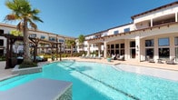 This single-story residence is part of a well-appointed community characterized by Mediterranean-inspired architecture, featuring a striking white stucco exterior with terracotta-accented rooflines and pergola-shaded courtyards. The centerpiece of the community amenities is a generously sized resort-style pool surrounded by lounge seating, lush landscaping, and an expansive covered patio—ideal for outdoor relaxation.

Inside, the open-concept floor plan seamlessly connects the living, dining, and kitchen areas into a cohesive, flowing space. Rich hardwood flooring runs throughout, lending warmth and continuity to the interior. The living area is well-lit by multiple windows dressed in classic plantation shutters and linen drapes, with a ceiling fan adding both style and comfort overhead.

The kitchen is finished with dark espresso cabinetry, granite countertops, stainless steel appliances, and a breakfast bar with pendant lighting—making it both functional and visually striking. Adjacent to the kitchen, the dining area features a chandelier overhead, providing an inviting setting for entertaining.

The entryway is accented with decorative wall art and a console table, creating a welcoming first impression. Throughout the residence, neutral tones and thoughtful design details contribute to a comfortable and cohesive aesthetic.