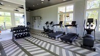 This fitness center offers a comprehensive workout environment designed to accommodate a wide range of exercise routines. The space is anchored by a row of modern treadmills and upright stationary bikes, providing ample cardio options for residents or members. Along one wall, a dedicated free weight station features a full dumbbell rack, while a TRX suspension training system is mounted nearby, supporting functional and bodyweight training exercises.

The interior design is both clean and contemporary, with light gray walls that create a calm, focused atmosphere. One of the most striking features of the space is the geometric patterned flooring, rendered in alternating tones of gray and white, which adds a bold visual element and a sense of energy to the room. Recessed lighting and ceiling fans keep the space well-lit and comfortable during intense workouts.

Large windows along the exterior wall flood the room with natural light and offer views of the surrounding landscaping and architecture, contributing to an open and inviting ambiance. The overall layout is thoughtfully organized, maximizing usable floor space while keeping equipment accessible and the environment uncluttered. This fitness center is well-suited for both light stretching sessions and more rigorous training routines.