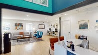 The Aspen Apartments leasing office, located at 4615 S Virginia St in Amarillo, TX, is a thoughtfully designed single-floor space that makes a striking first impression. The reception area anchors the entrance with a sleek white desk, cognac leather guest chairs, and bold teal and navy accent walls that rise to dramatic double-height ceilings adorned with clerestory windows, flooding the space with natural light. Light hardwood flooring runs throughout, unifying the open layout. Adjacent to the reception desk, a comfortable lounge area features a contemporary gray sofa, a pair of vibrant teal accent chairs, a wood-and-metal coffee table, and colorful wall art — all arranged on a layered area rug that adds warmth and texture. The business center, seamlessly integrated into the open floor plan, provides workstations equipped with desktop computers, gold-accented desk lamps, and ergonomic seating. A dedicated refreshment station with white shaker cabinetry, granite countertops, a beverage cooler, and a curated snack display offers a welcoming amenity for residents and guests alike. Recessed lighting throughout enhances the bright, airy ambiance, while multiple windows provide outdoor views and additional natural illumination. The overall design balances professionalism with comfort, creating an inviting and functional community hub.